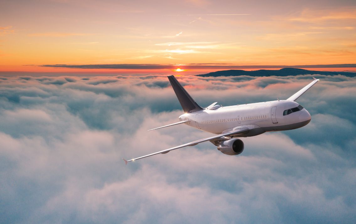 Large white airplane flying above white/blue/grey clouds with sunsetting in background with orange, yellow, purple sky