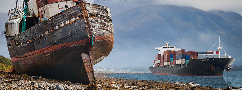 Old broken wooden ship beached on the sand while a new cargo ship sits in the water